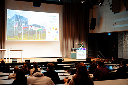 Dr. Alain Bécoulet, Deputy Director at ITER Organization, presents on the future of fusion energy at binding.energy, pointing to a slide of the ITER main building with international flags while speaking on the main stage before a focused audience