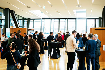 Participants at binding.energy nuclear conference exchanging ideas during a coffee break between fusion and fission presentations, gathered in small groups at standing tables with drinks and snacks