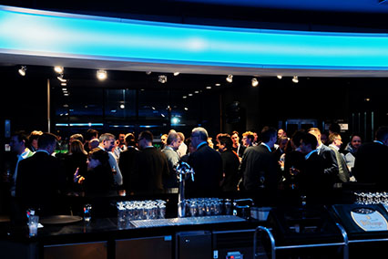 Evening networking atmosphere at binding.energy in the iconic Tivoli Stadium bar, with fusion and fission experts, speakers, and participants gathered in lively conversation under futuristic blue lighting