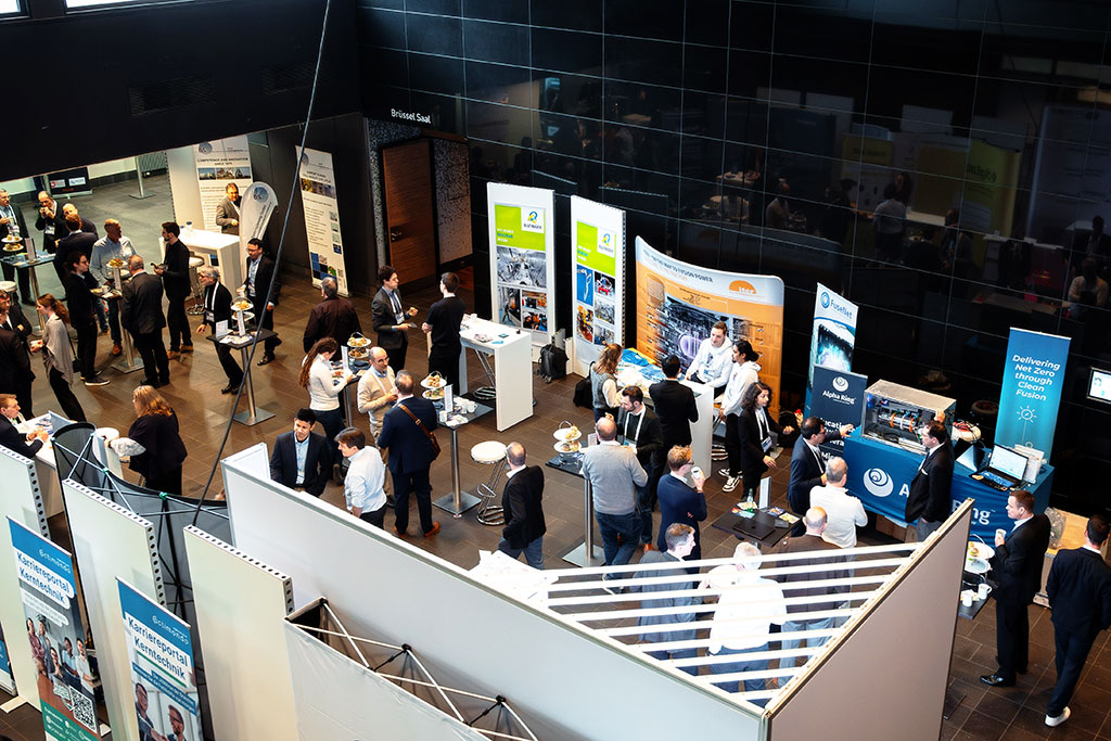 Nuclear industry professionals engaging at sponsor booths in the exhibition hall of binding.energy, featuring brands like Alpha Ring, ITER, Bilfinger, Brenk Systemplanung, and organizer actimondo eG.