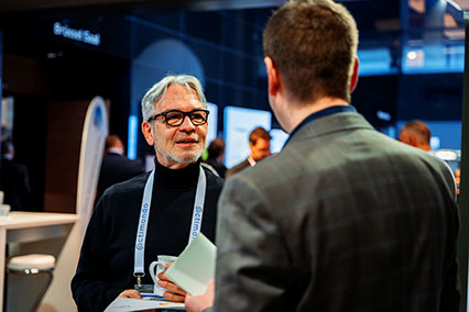 Two nuclear experts in formal attire engaged in conversation at the binding.energy exhibition hall; one facing the camera with white beard and glasses, the other holding documents — reflecting the professional tone of the conference