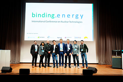 The organizing team of binding.energy poses on the main stage in front of the official conference banner and sponsor logos — representing the dedicated people who brought the international fusion and fission event to life