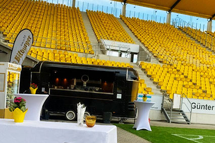 Binding.energy participants are welcomed with warm food and cold drinks at the Tivoli stadium, featuring a grill stand, drink station, and decorated tables on the pitch, with a view of the stadium stands in the background