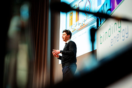 Tobias Ravn Thomsen from Copenhagen Atomics delivering a captivating talk on mass manufacturing thorium reactors during binding.energy, photographed through a microphone silhouette with the conference logo clearly visible at the speaker’s podium