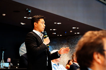 Young international nuclear expert in suit engages in the binding.energy conference by asking a pointed question on fusion-fission synergies during Topic 3 discussion